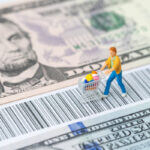 A miniature consumer figurine with a shopping cart walking on the bar code with us dollar banknotes money