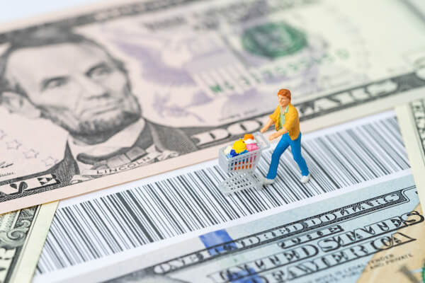 A miniature consumer figurine with a shopping cart walking on the bar code with us dollar banknotes money