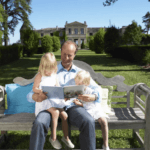 A rich American reading a book with his daughters on a bench in front os their detached house