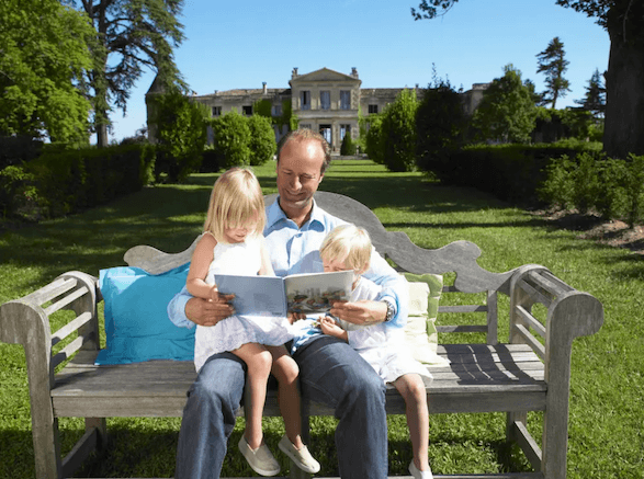 A rich American reading a book with his daughters on a bench in front os their detached house