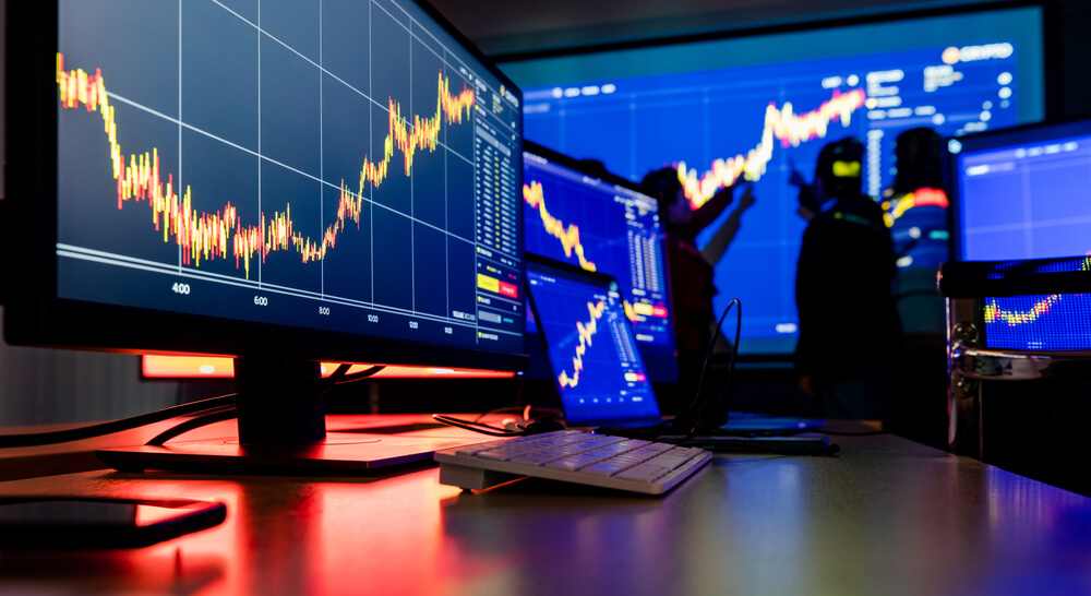 Multiple computer monitors displaying colorful stock market charts and trading data in a dark, high-tech environment, with people analyzing the projected graphs on a large screen in the background.