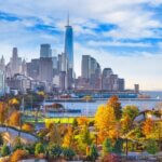 New York City skyline with One World Trade Center rising above the buildings, viewed from a park with colorful autumn trees and the Hudson River in the foreground.