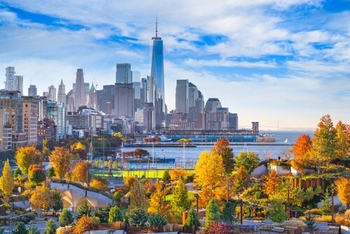 New York City skyline with One World Trade Center rising above the buildings, viewed from a park with colorful autumn trees and the Hudson River in the foreground.