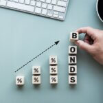 An overhead view of a desk where a hand stacks wooden blocks to spell the word 'BONDS' alongside blocks with percentage signs, creating an upward-trending financial graph to symbolize rising bond yields and interest rates.