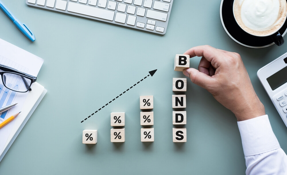 An overhead view of a desk where a hand stacks wooden blocks to spell the word 'BONDS' alongside blocks with percentage signs, creating an upward-trending financial graph to symbolize rising bond yields and interest rates.