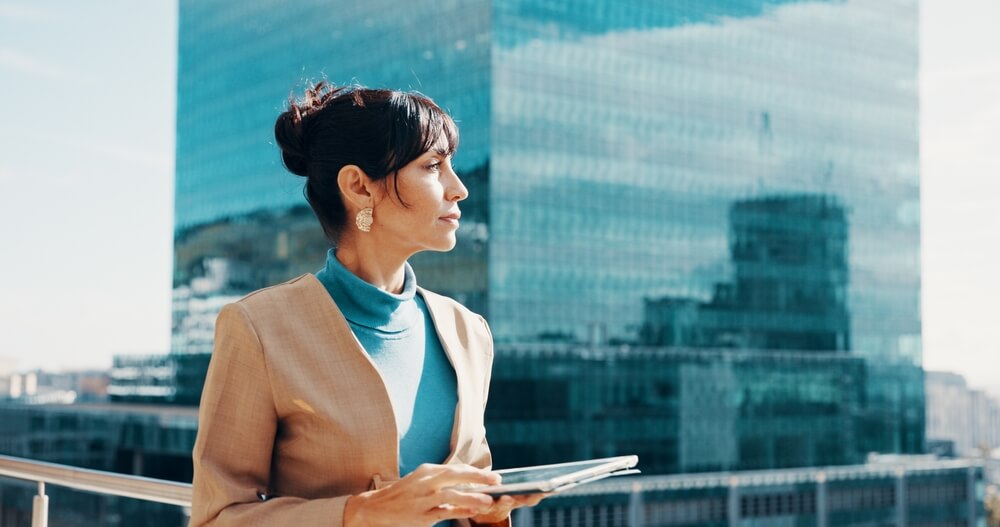 A professional woman holding a tablet and looking thoughtfully toward the horizon outside a modern glass office building, symbolizing a forward-looking, prepared approach to long-term wealth management.